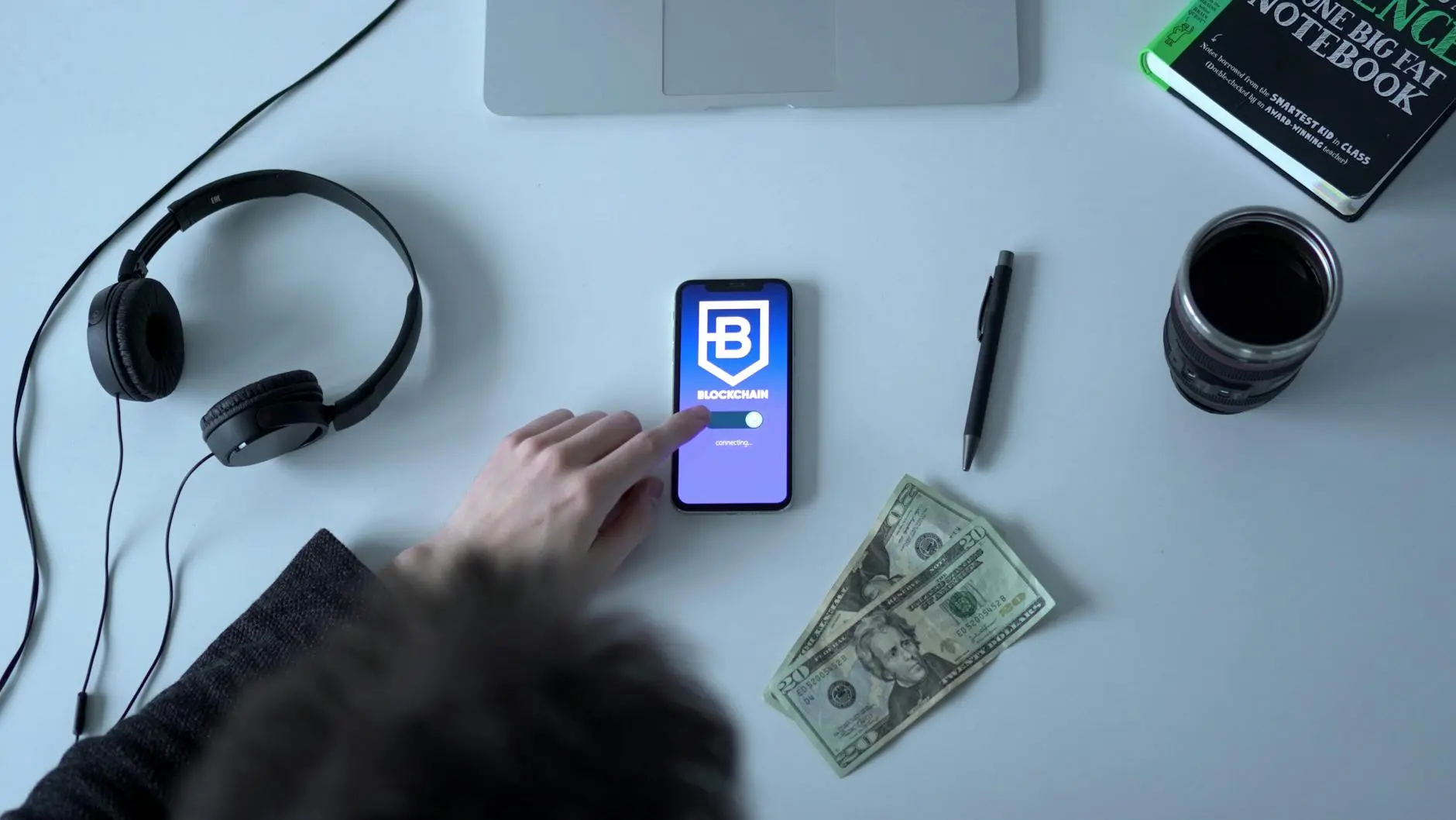 A smartphone displaying a blockchain app, headphones, cash, and a notebook on a desk.