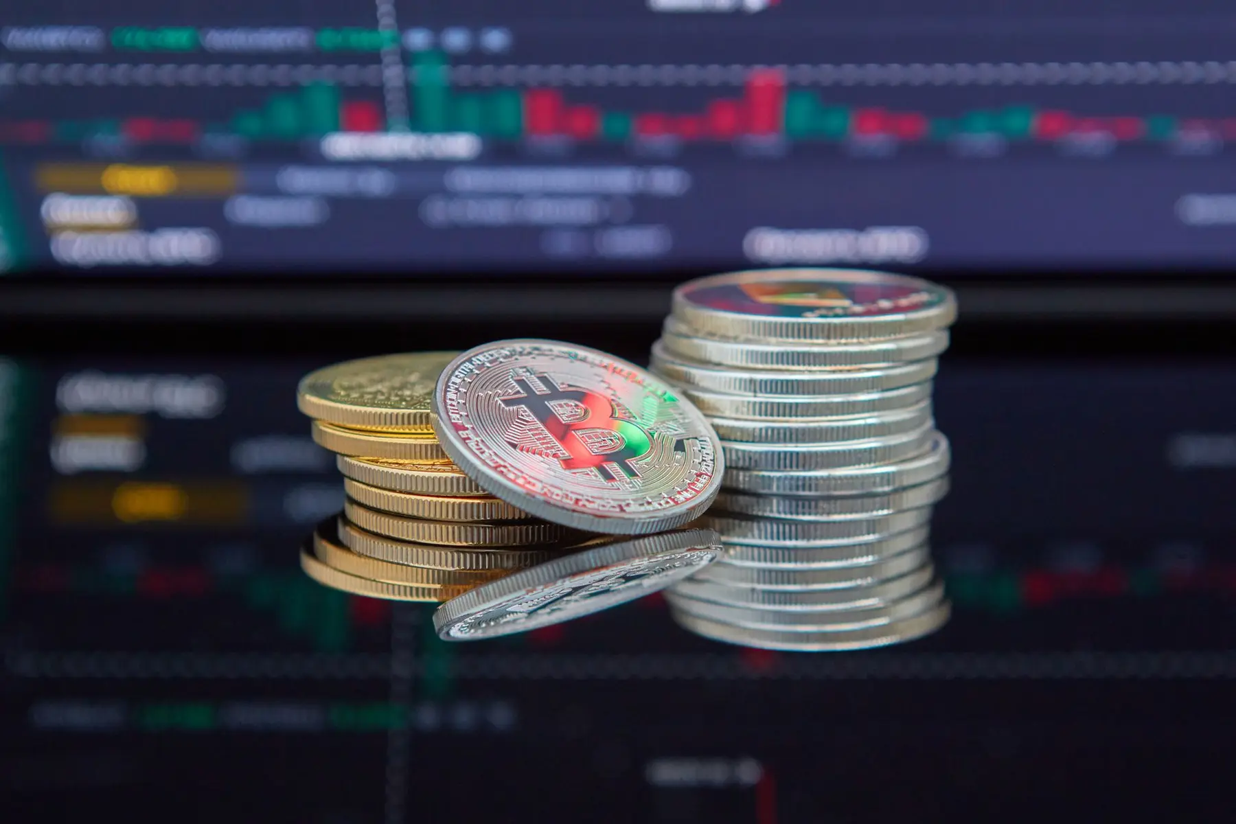 Physical Bitcoin and silver cryptocurrency coins stacked in front of a blurred digital trading chart