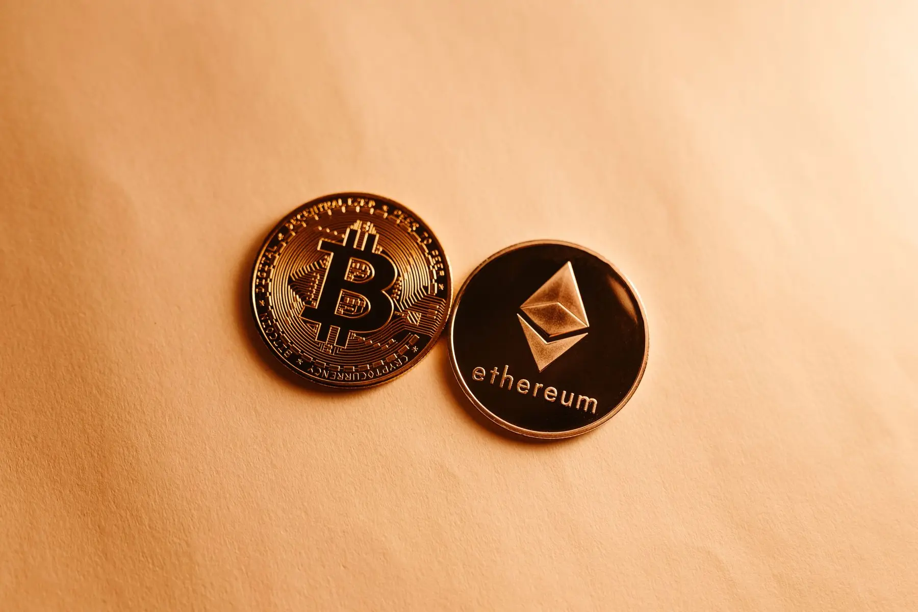 Physical gold Bitcoin and silver Ethereum cryptocurrency tokens resting on a plain tan surface