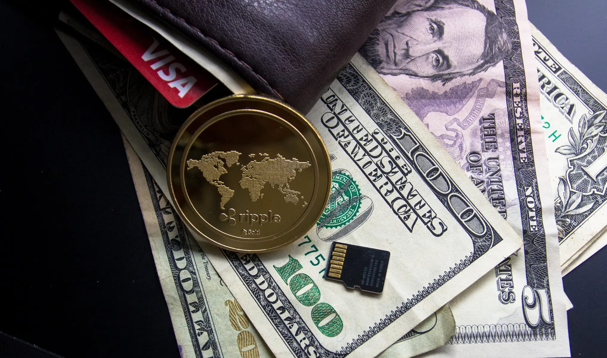 A gold physical Ripple XRP coin resting on US dollar bills, a wallet, and a credit card.