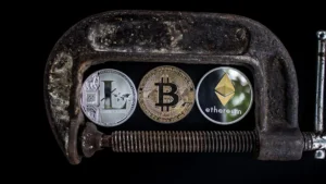 Physical cryptocurrency coins including Bitcoin and Ethereum held under a metal C-clamp against a black background.